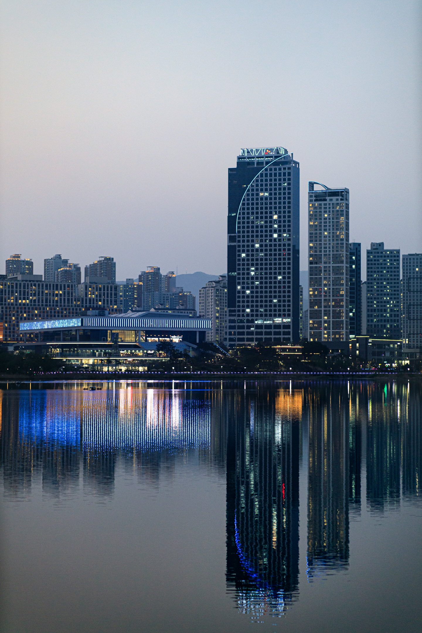 Business and apartment buildings stand at twilight over their blurred reflections in the ripples of a lake. Neon lights, barely visible on the towers, are revealed in the dark waters.