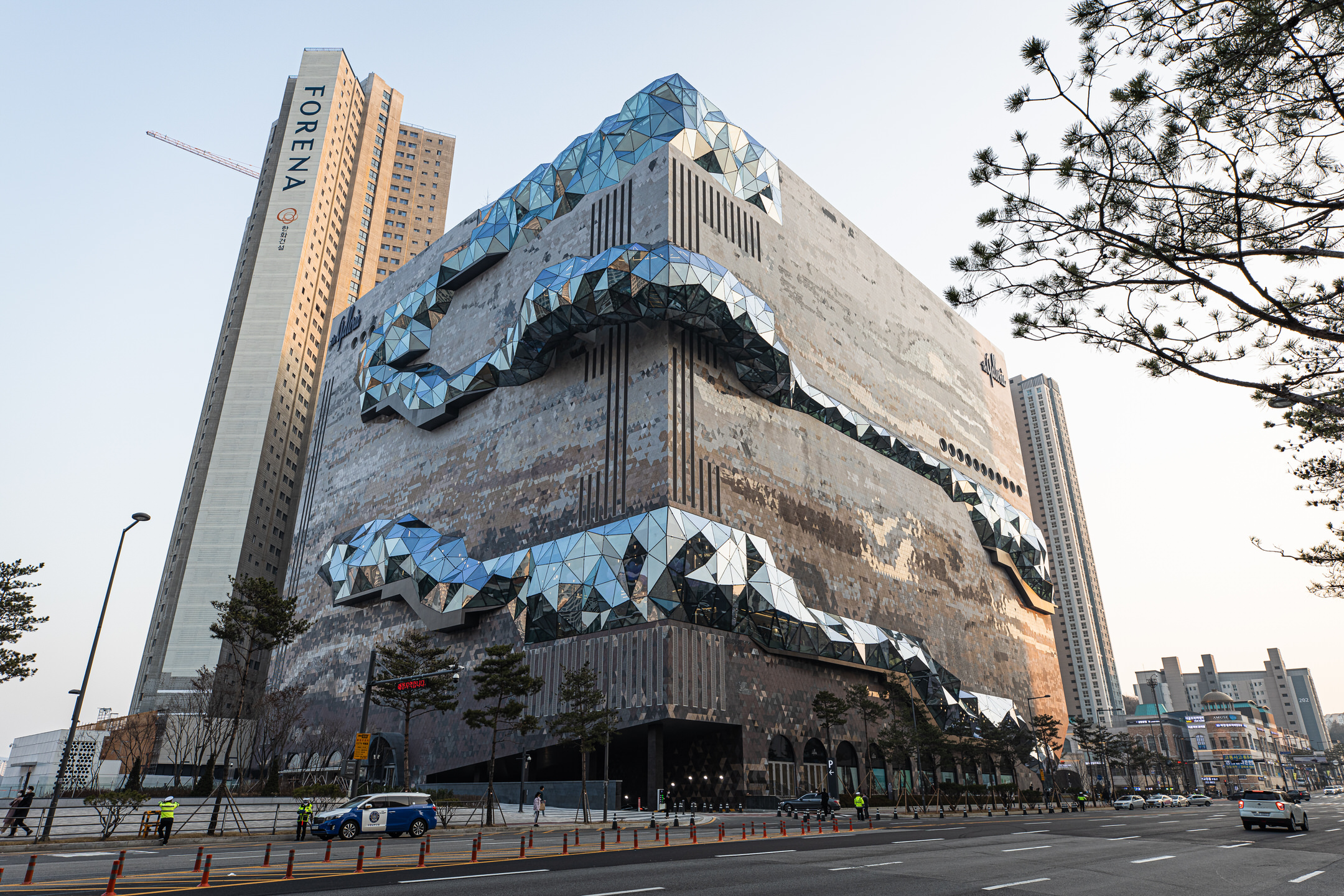 Full-shot of a cubic department store from the street. The matte exterior of the building shows darker, brownish colors at the bottom that progressively and erratically morph into lighter, grayish layers as we progress to the top. Between those layers, a serpentine vein-like growth mars the surface of the walls with a low-polygon count, triangular, silvery window surface that spirals to the top.